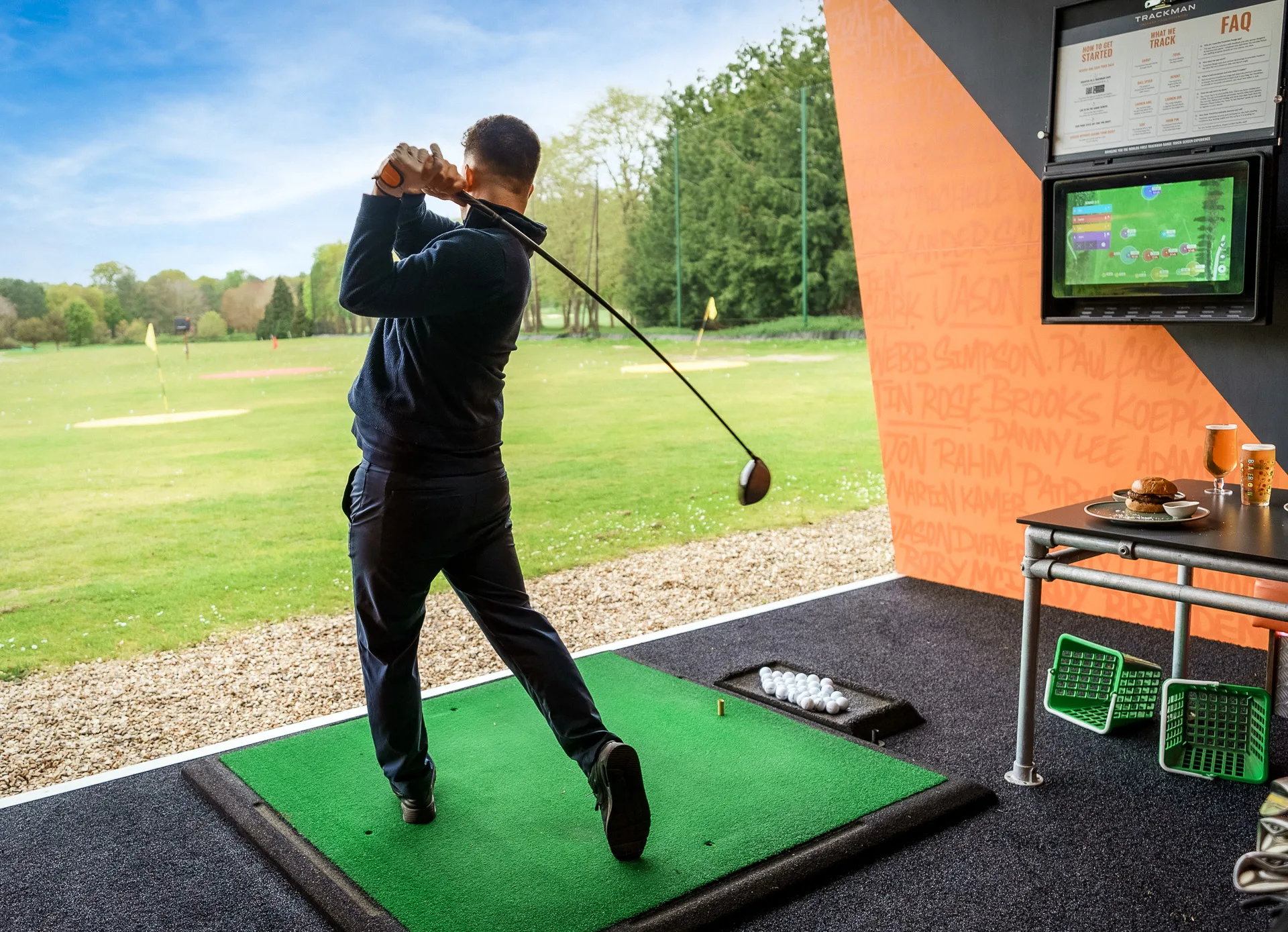 A man swinging a golf club in woking, hitting a ball out of a driving range bay, with the ball in mid-flight and the range stretching out ahead under an open sky