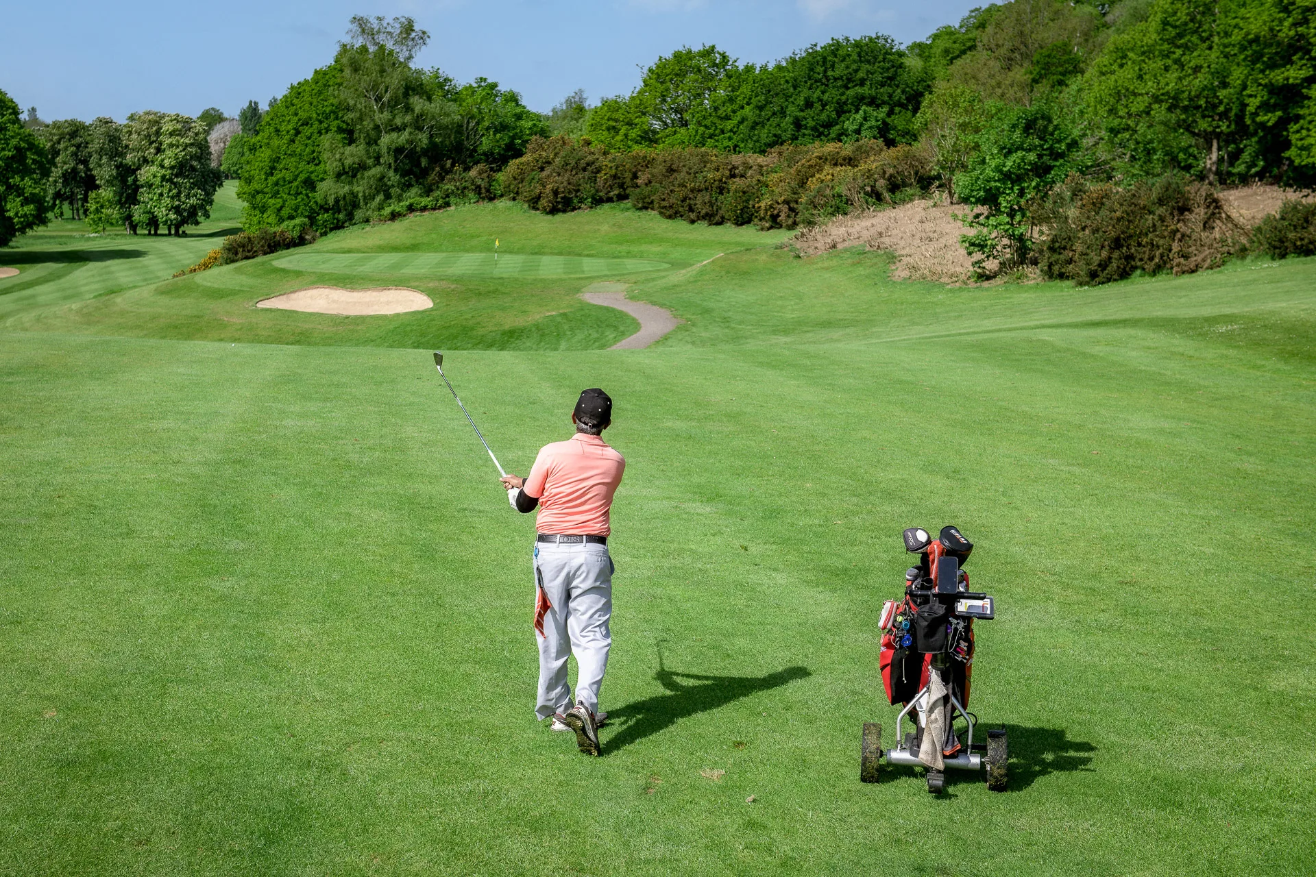 Golfer watching ball flight towards the green with a trolley on the fairway at a golf course in Woking