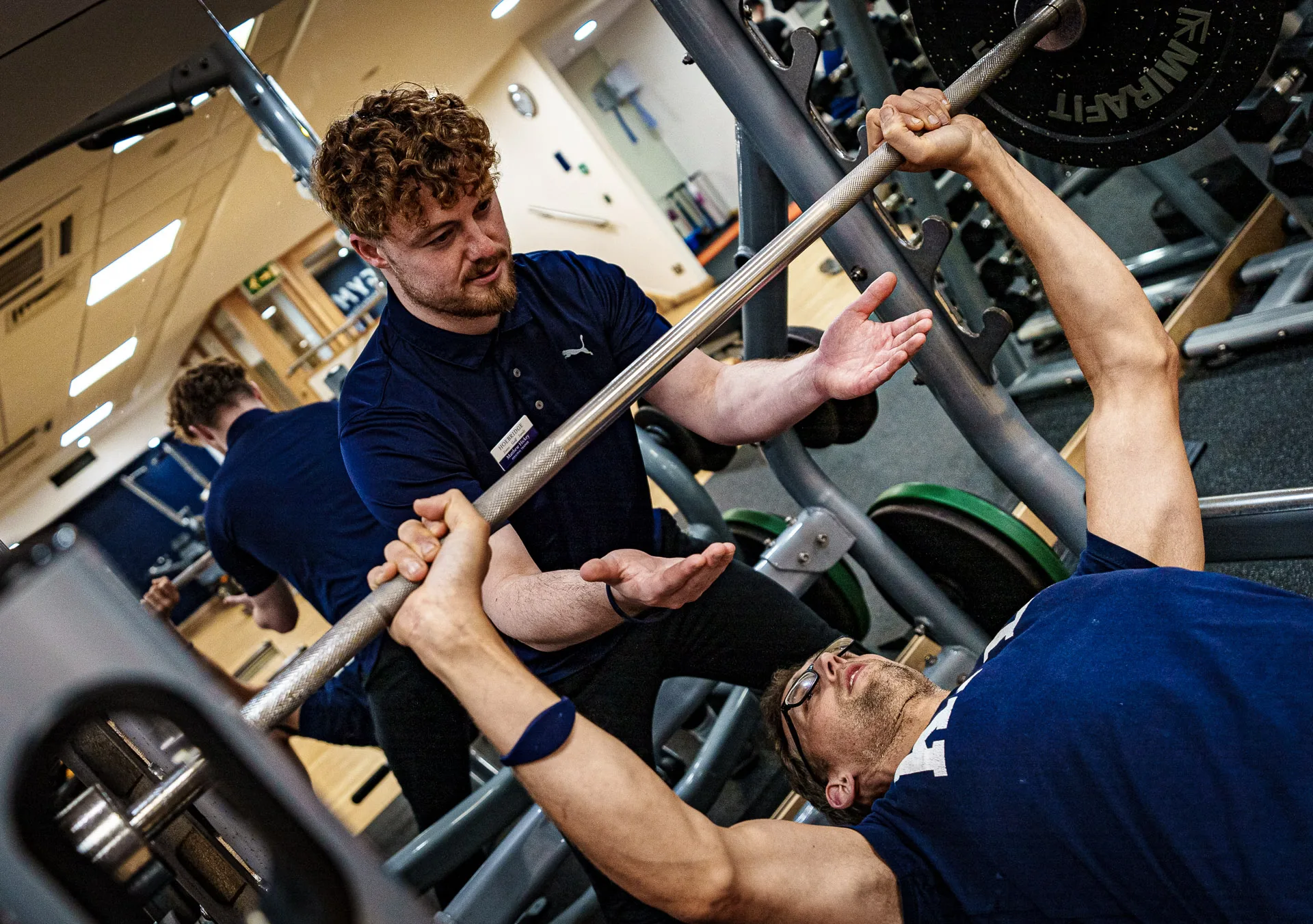 A personal trainer at a gym in woking closely spotting a client during a bench press in the gym, ensuring proper form and safety. The client is lying on a bench with a barbell, focused and determined